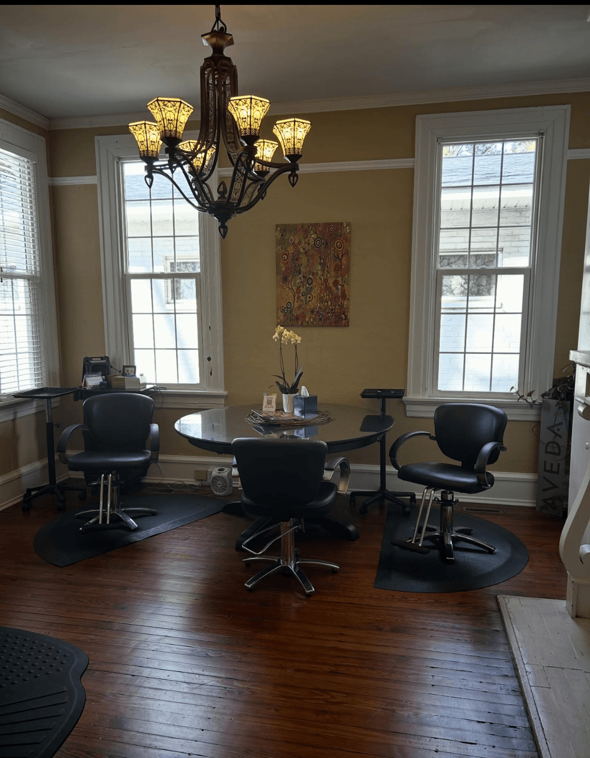 Dining room with a round table, four chairs, a chandelier, and three large windows letting in natural light.