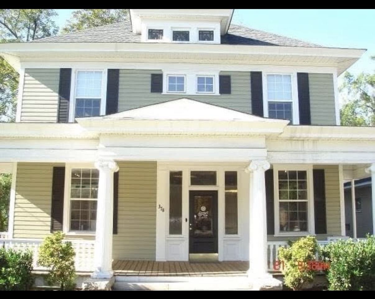 Front view of a two-story house with a porch, columns, and multiple windows, surrounded by greenery.