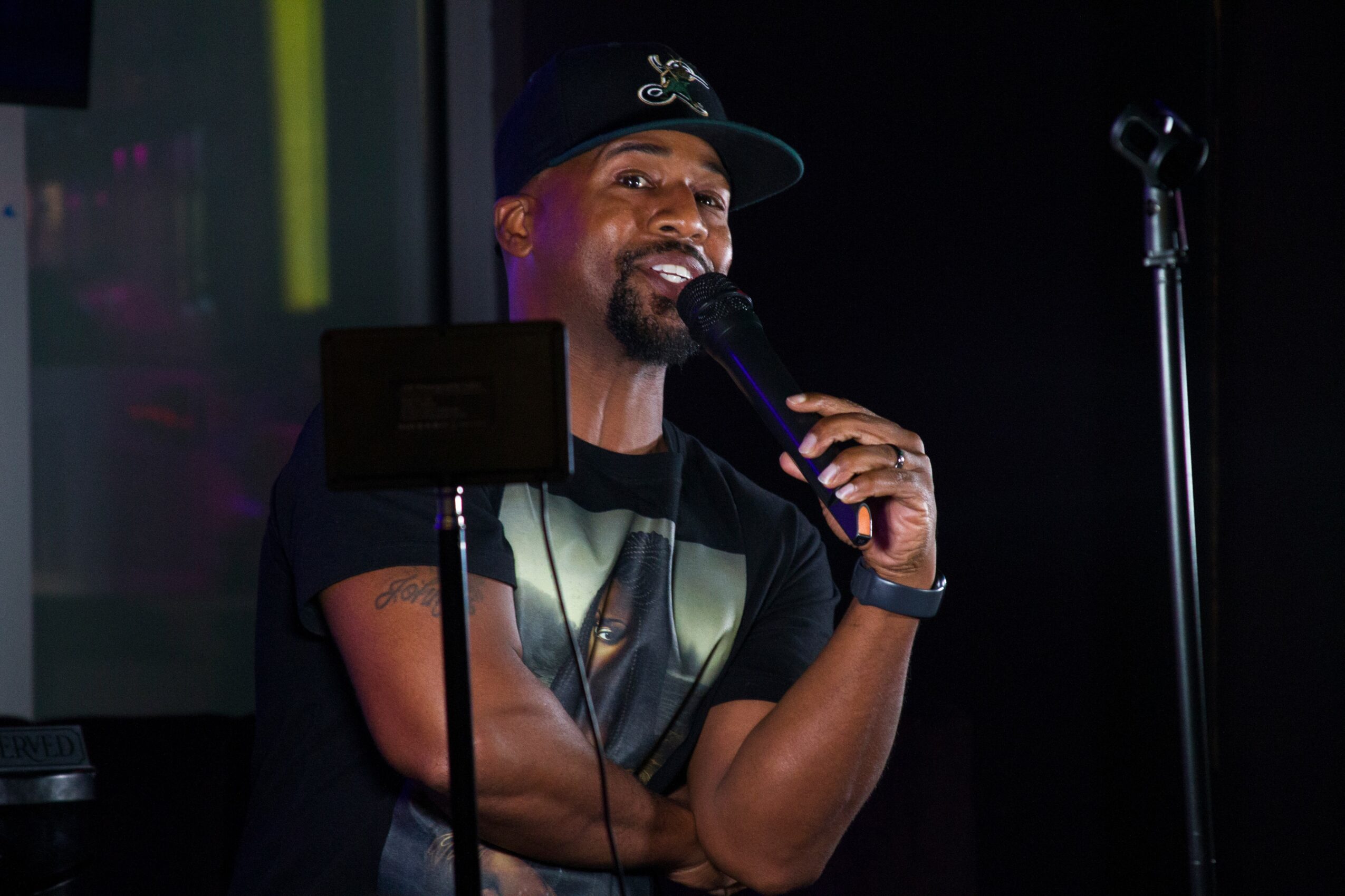 Person smiling, holding microphone, wearing a cap and black T-shirt, on stage with dark background.