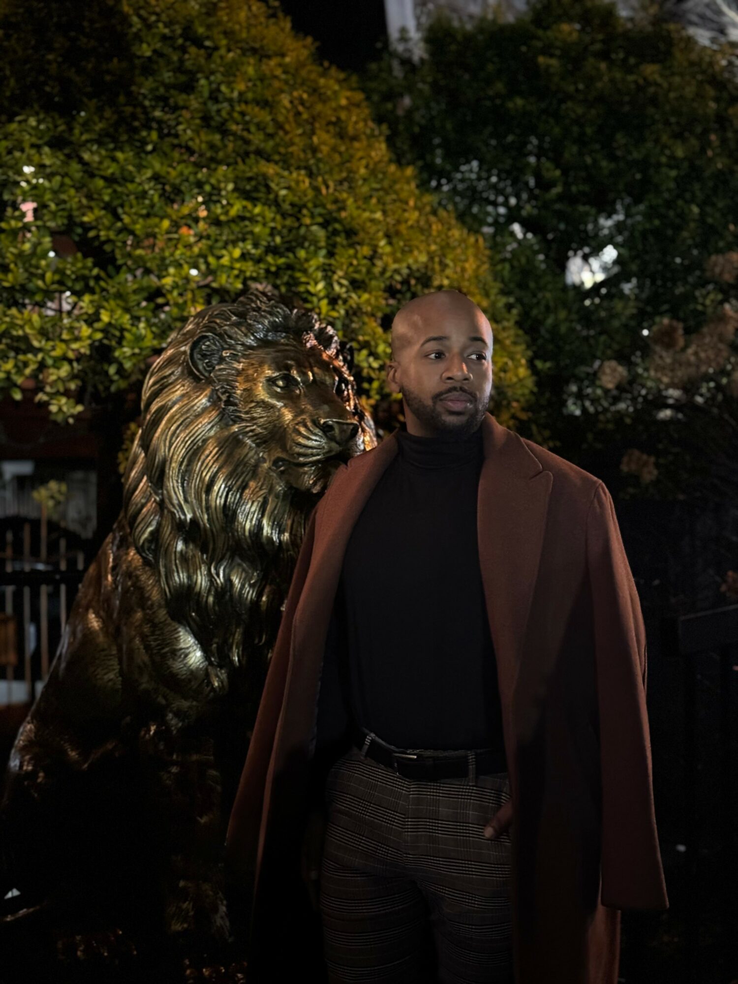 Man standing outdoors with a lion statue, trees in the background, evening lighting, wearing a brown coat and black shirt.