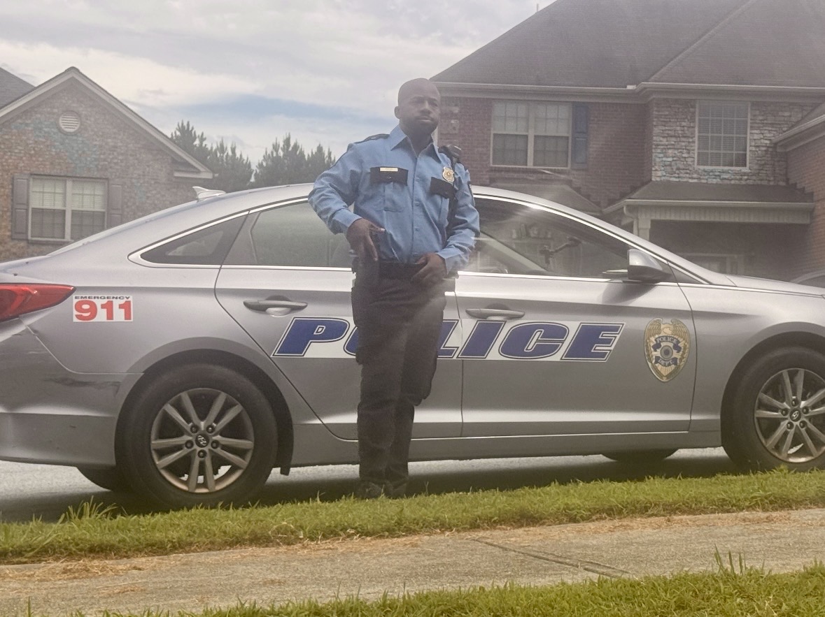 Police officer standing next to a police car on a residential street, with houses in the background.