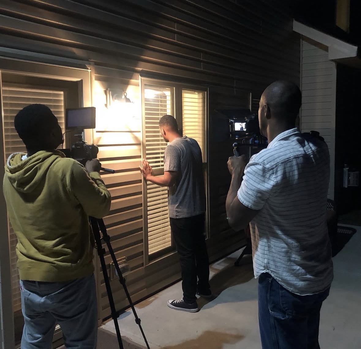 Three men filming a person writing on a wall with windows behind, at night. One man is using a camera on a tripod, another holds a camera, and the third writes on the wall.