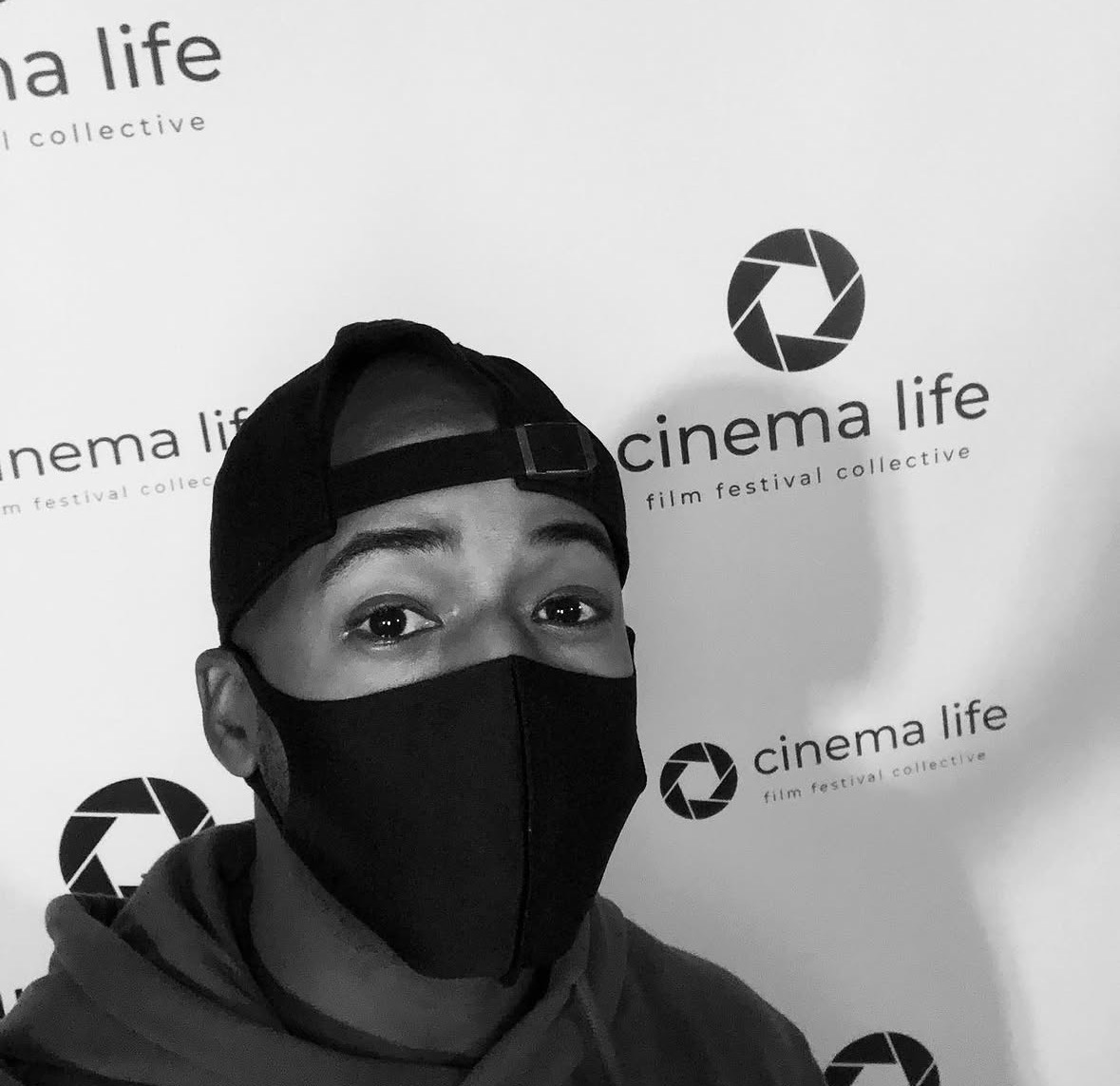 Person wearing a black face mask and cap, standing in front of a backdrop with 'cinema life' logos.