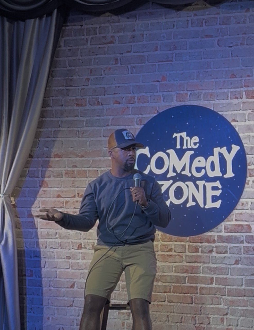 Person standing on stage with a microphone, gesturing with one hand, in front of a brick wall and a sign reading 'The Comedy Zone'.