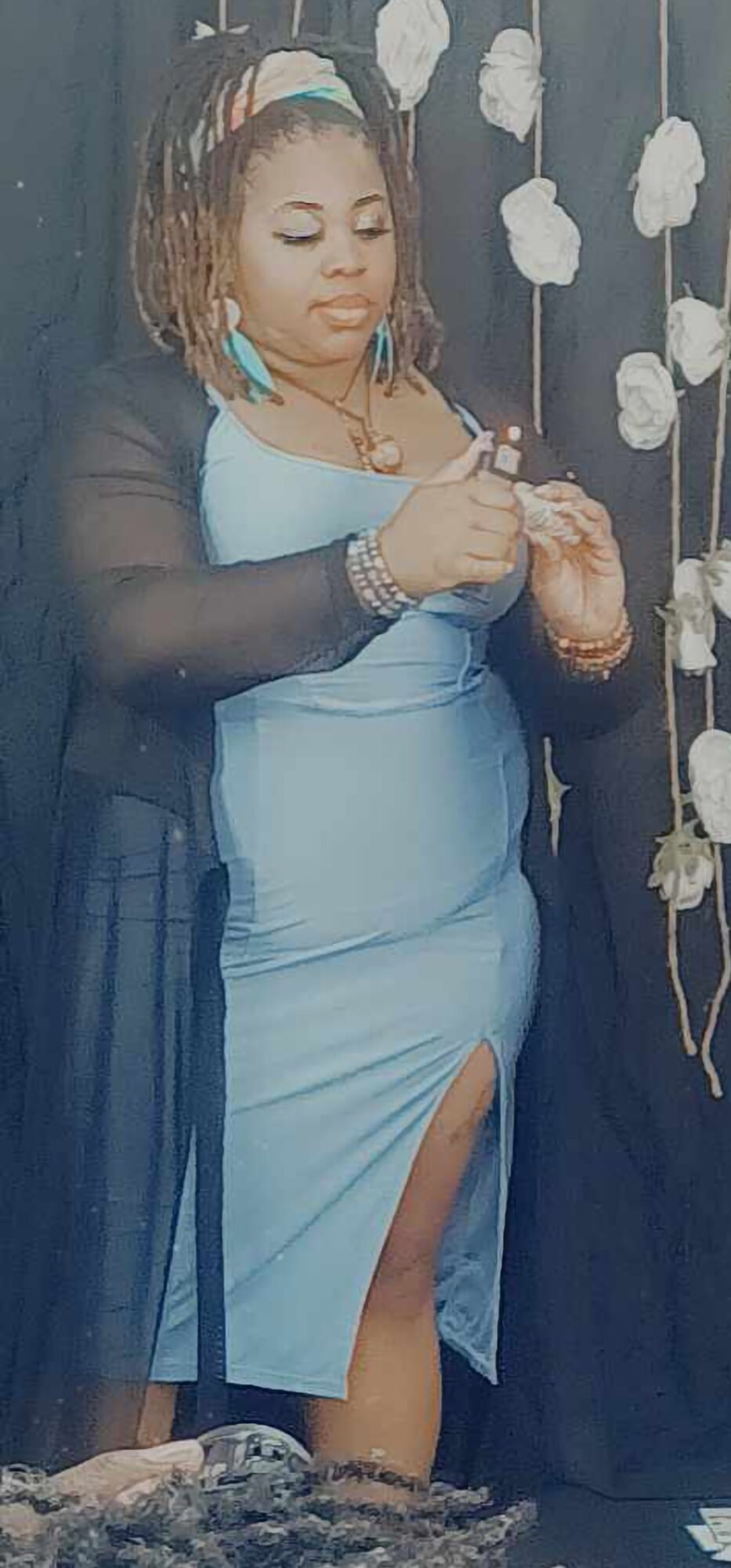 Woman in a light blue dress with a slit, standing near white flowers, looking down at her hands.