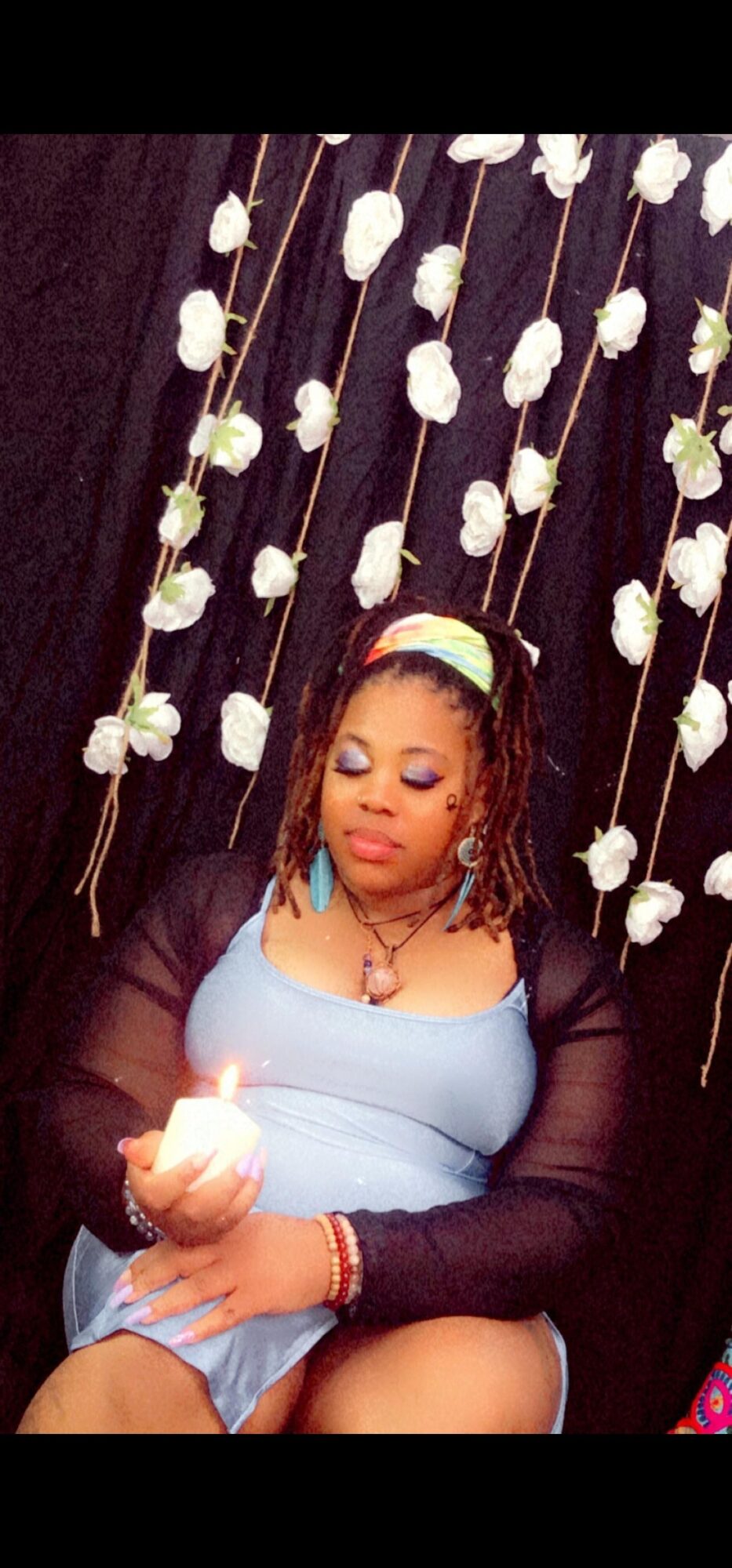 Woman with closed eyes holding a lit candle, sitting in front of a floral backdrop, wearing a light blue dress and black cardigan.