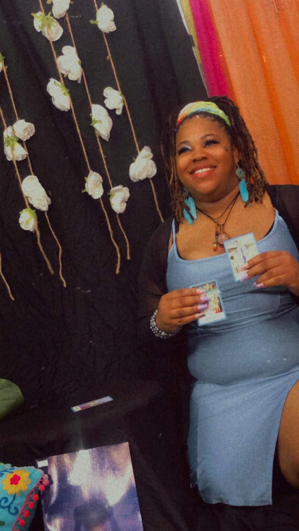 Smiling woman in blue dress holding a drink, decorated with flowers and colorful fabric backdrop.