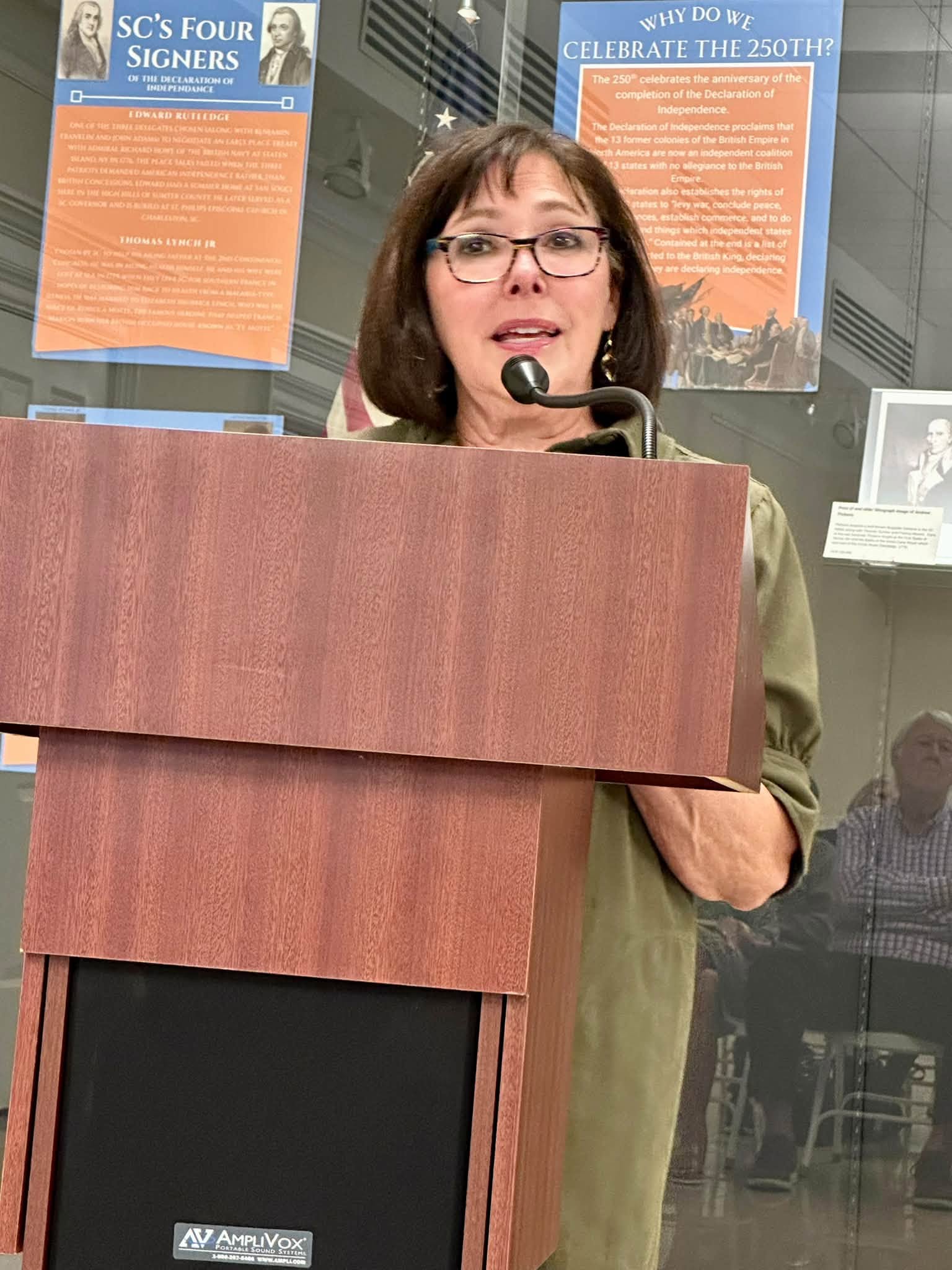 A person wearing glasses speaks into a microphone behind a wooden podium, with informational posters on the wall.