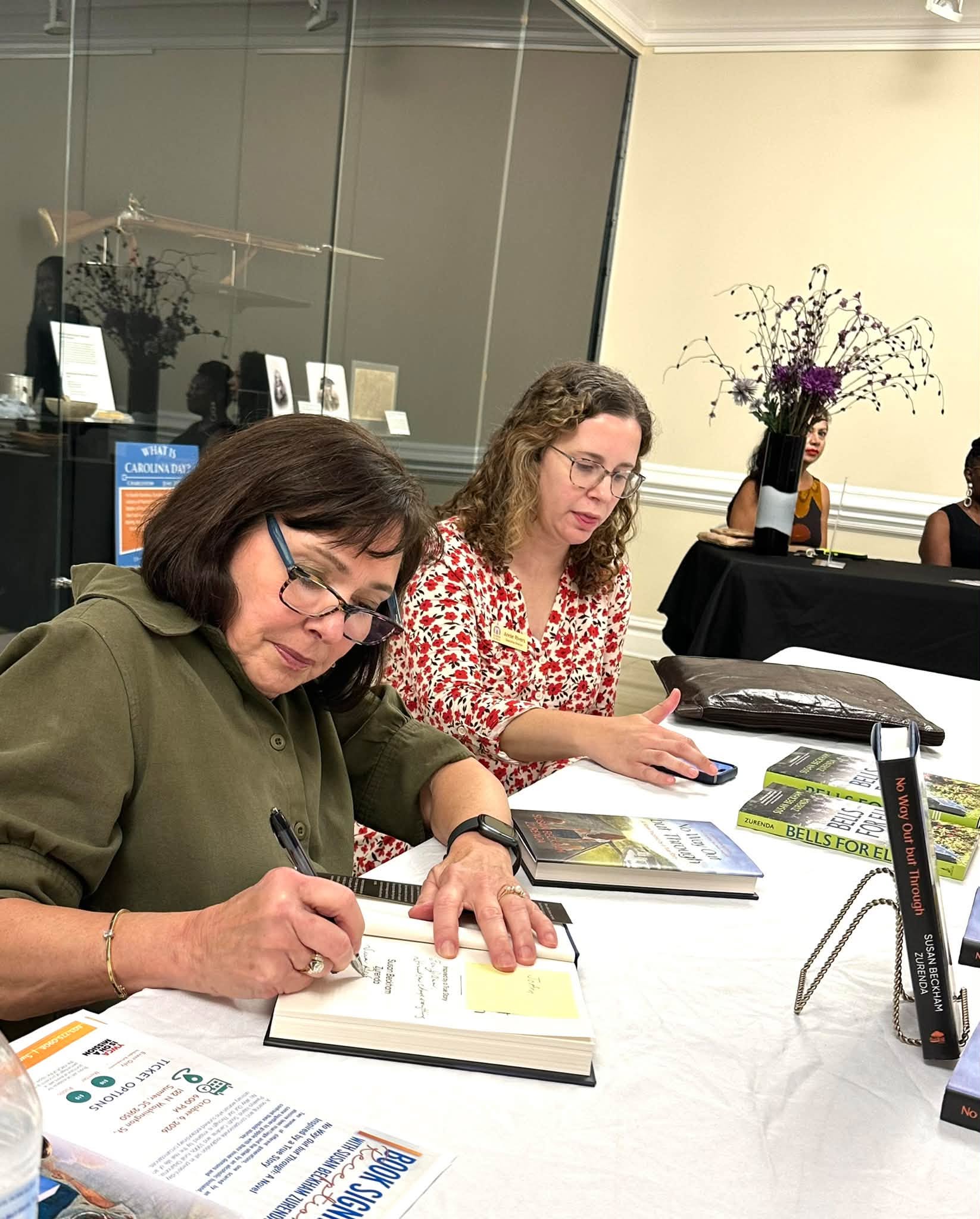 Two people sit at a table reviewing papers and writing, with a flower arrangement and display cases behind them.