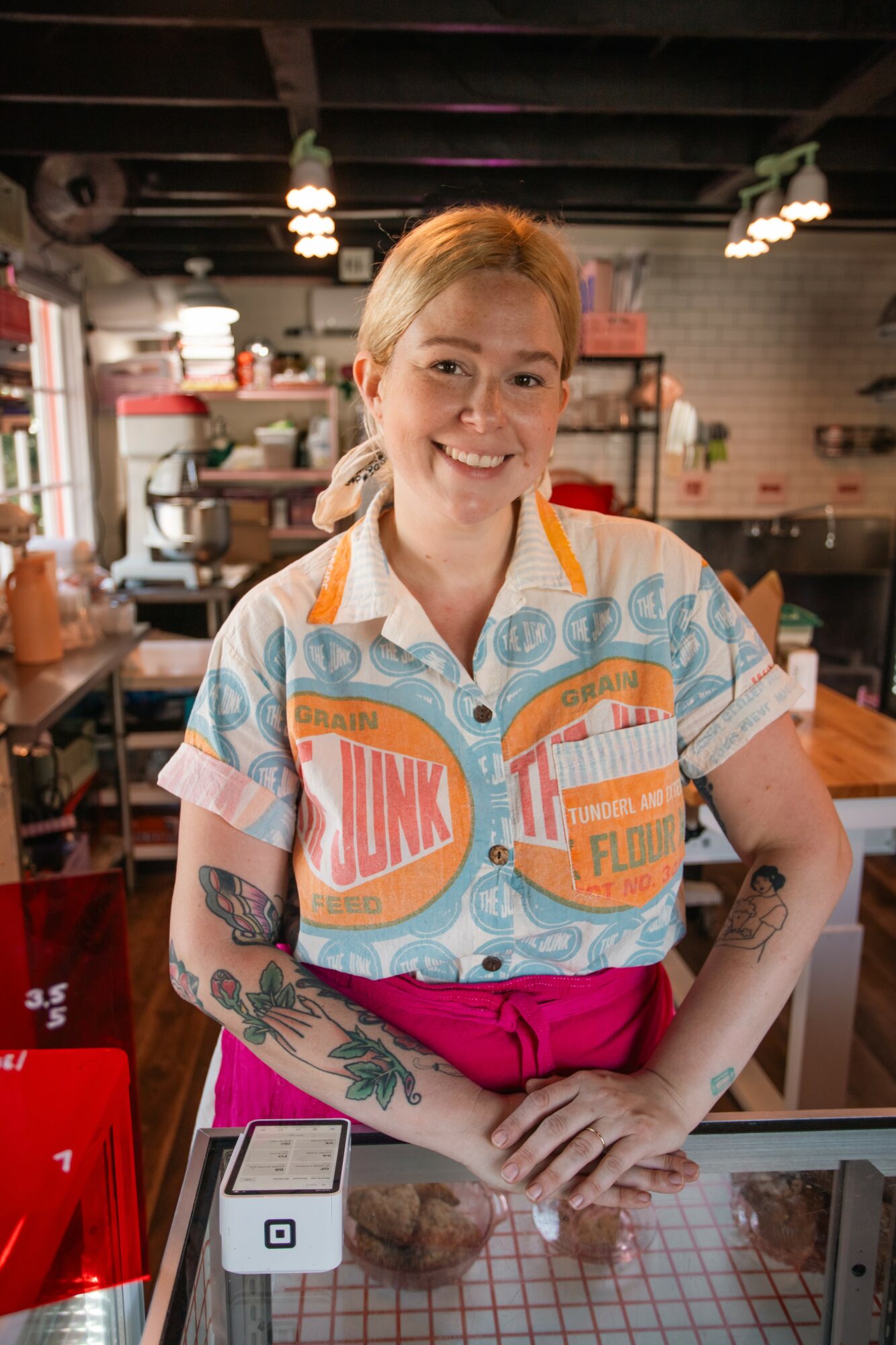 Young woman with blonde hair smiling, wearing a colorful shirt, standing in a bakery or cafe with shelves and baked goods in background.