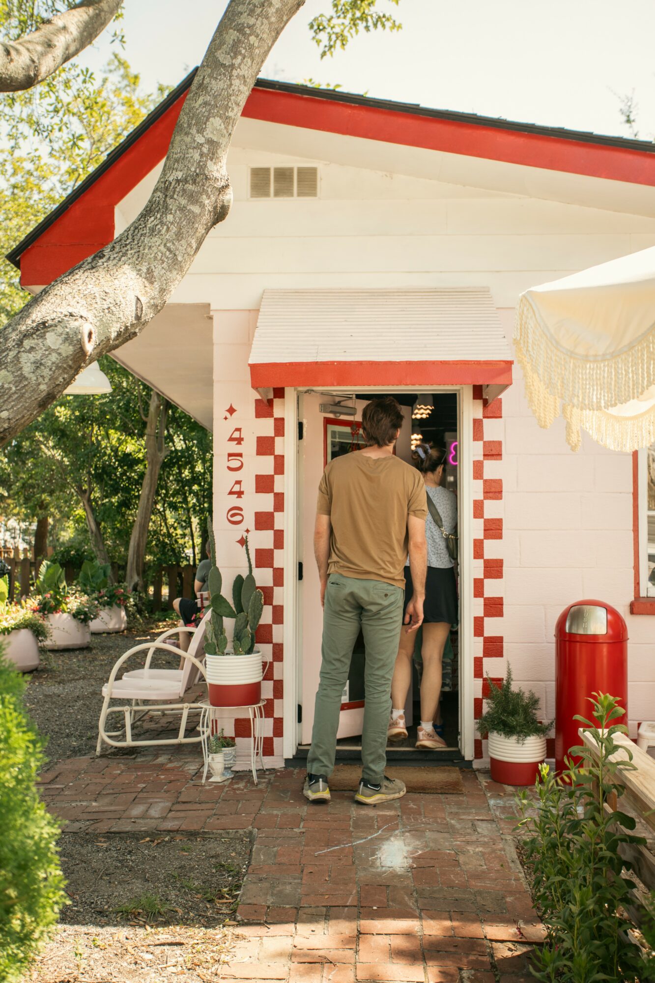 People entering a small building with red and white checkered trim, surrounded by outdoor furniture and greenery.