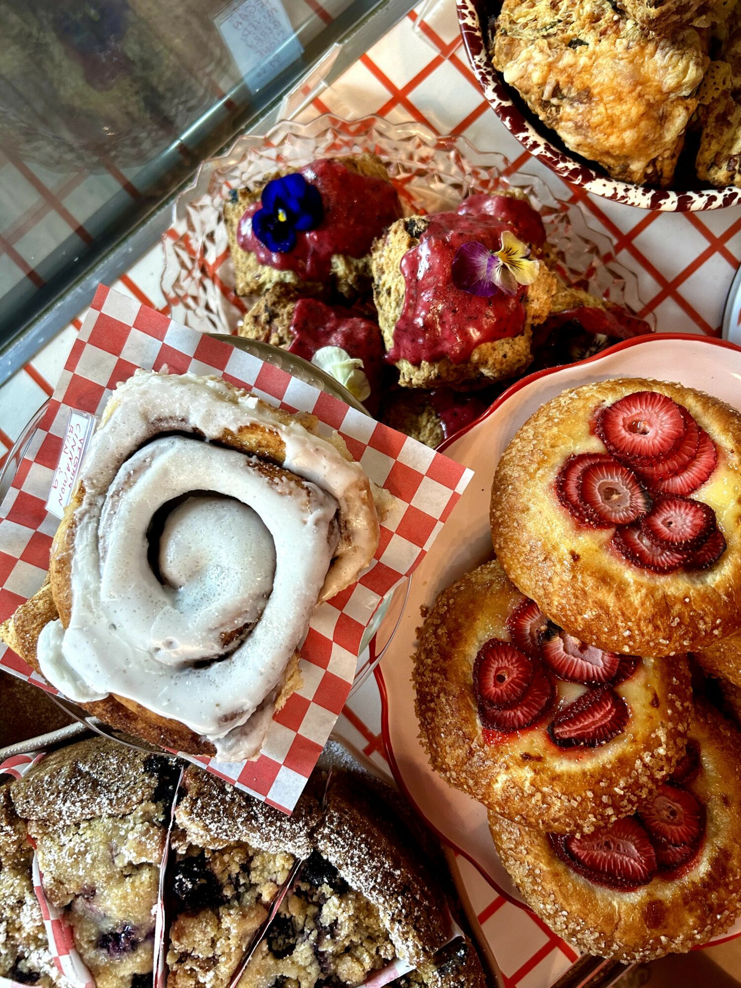 Assorted baked goods including cinnamon rolls with icing, berry-topped muffins, and a crumb cake with strawberries, on checkered paper