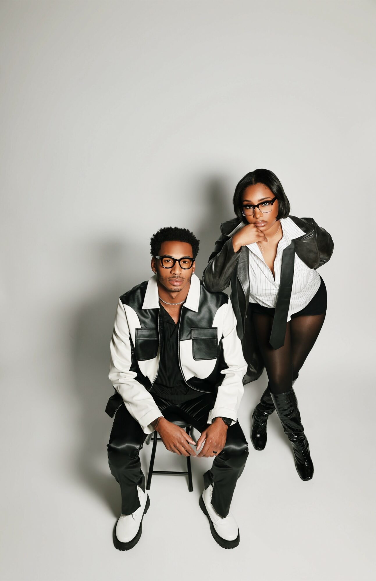 Two women with glasses and dark hair pose against a plain white background, one sitting and one standing, both dressed in black and white outfits.