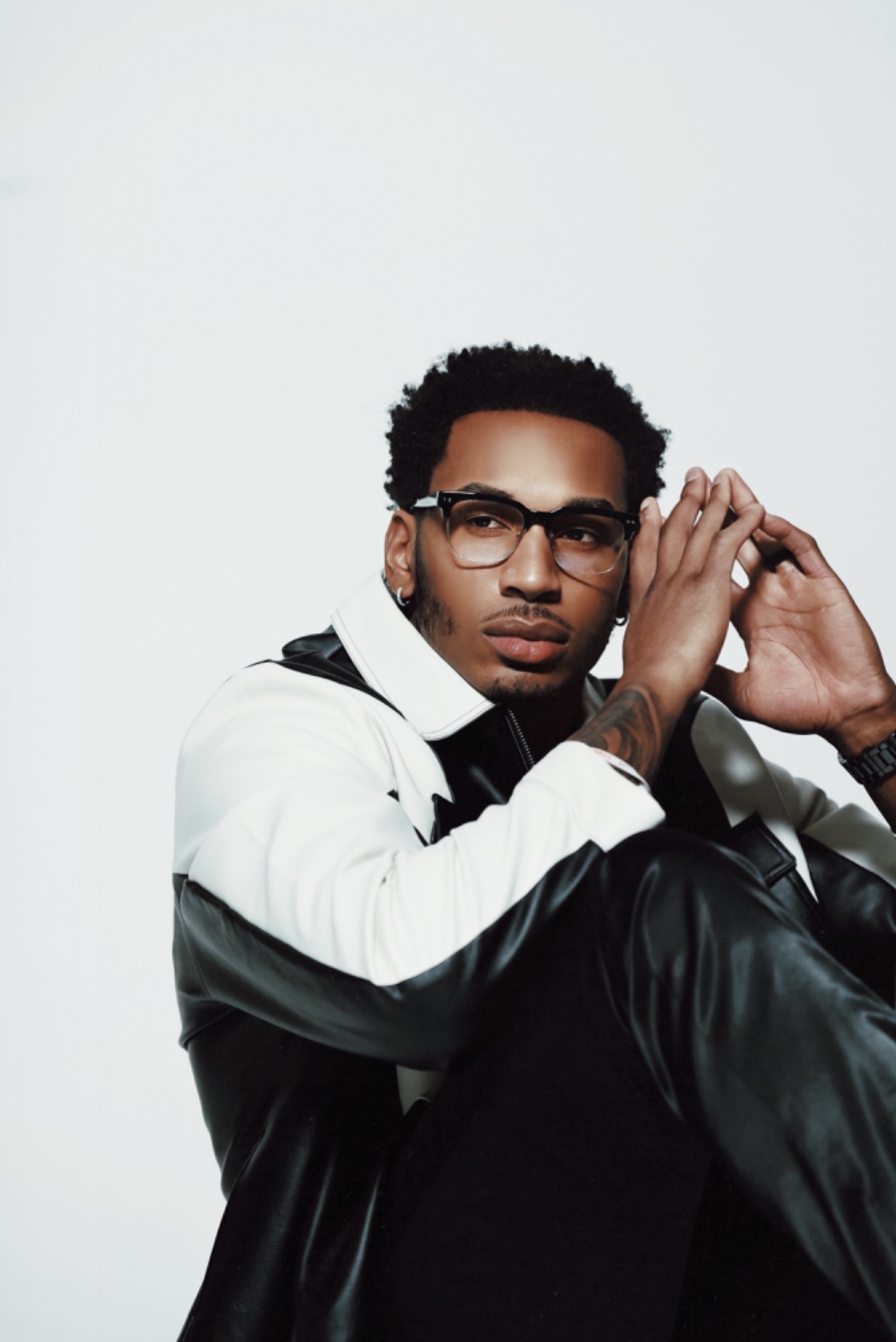 Young man with glasses and earrings, wearing a black and white jacket, sitting with hands near face, against a plain background.