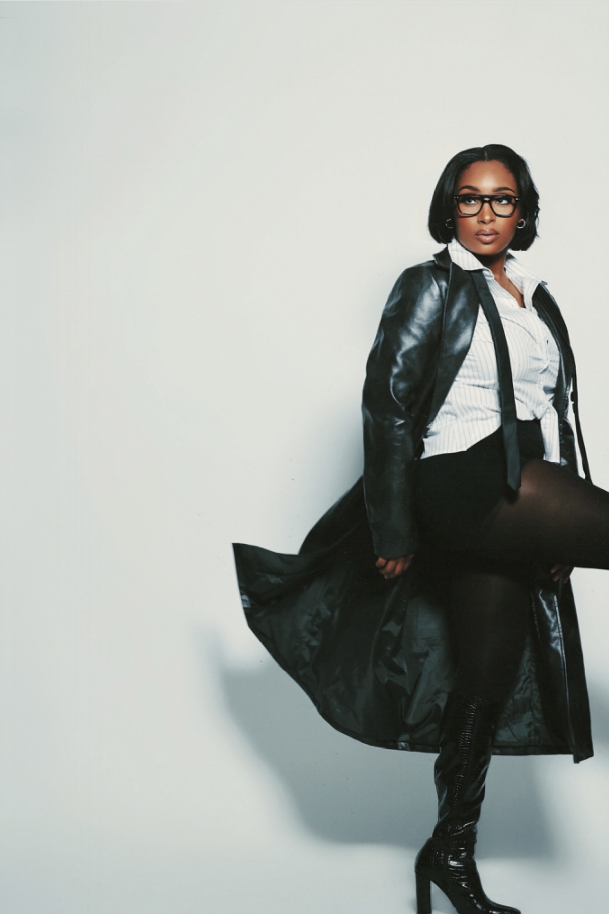 Woman with glasses in a leather jacket, white shirt, and black pants, standing against a plain light background.