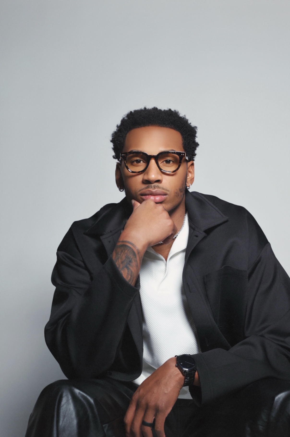 Man with glasses and curly hair, wearing a black jacket and white shirt, resting chin on hand, against a plain background.