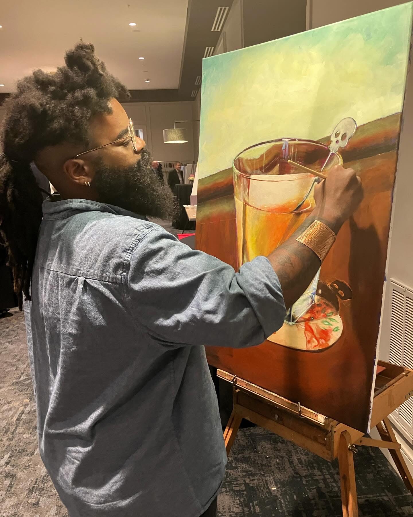Person with glasses and dreadlocks painting a still life with a glass, skull, and fruit on canvas.