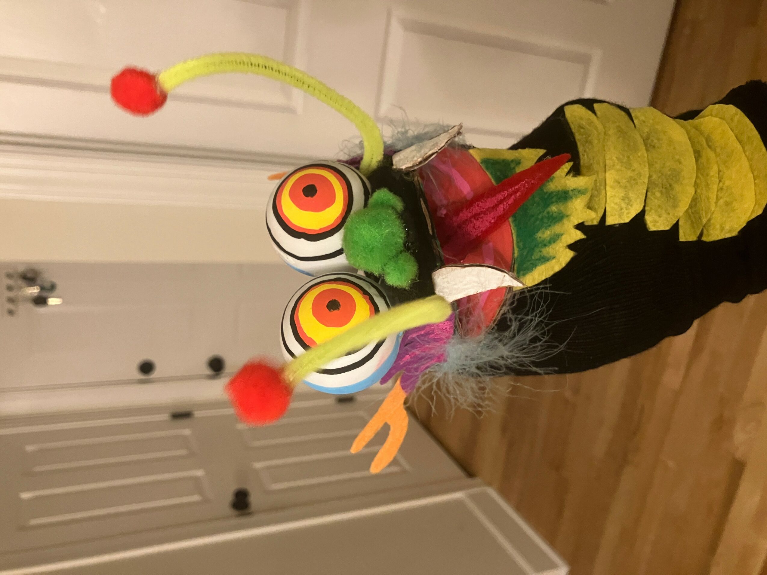 Colorful puppet with large eyes, antennae, and yellow and black striped body, standing on a wooden floor.