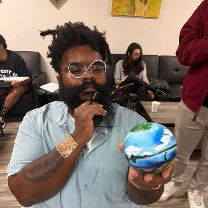 Man with glasses and beard holding a small globe, looking at it thoughtfully, in a room with people sitting on a couch.
