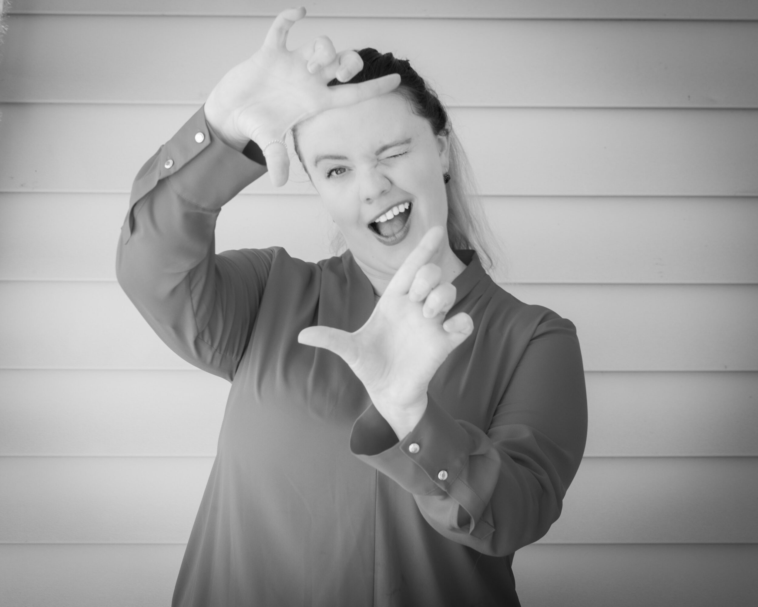 Woman making a playful gesture with hands, winking, and smiling, in front of horizontal siding, in black and white.