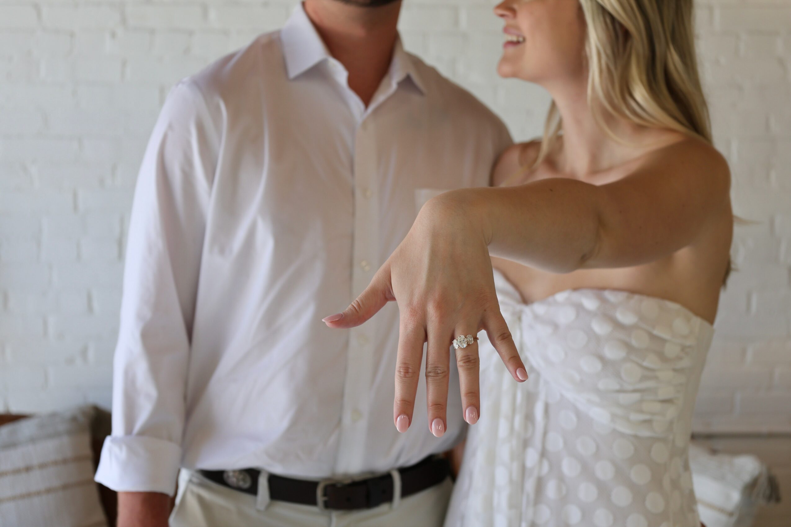 A woman shows her engagement ring on her finger, standing next to a man in a white shirt, smiling.