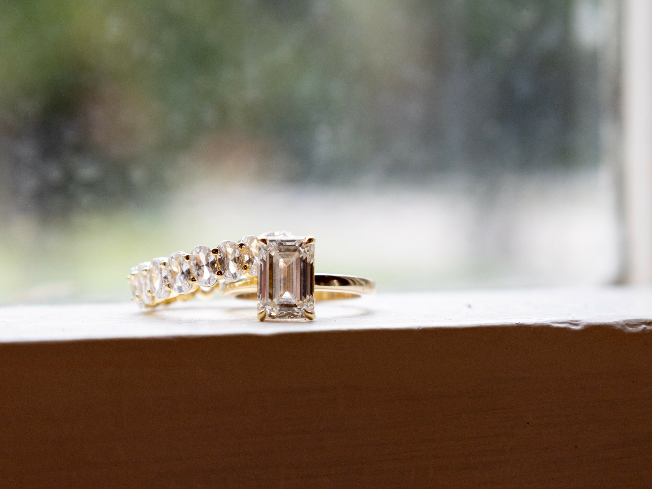 Ring with rectangular gemstone and small surrounding stones on a windowsill, blurred outdoor background
