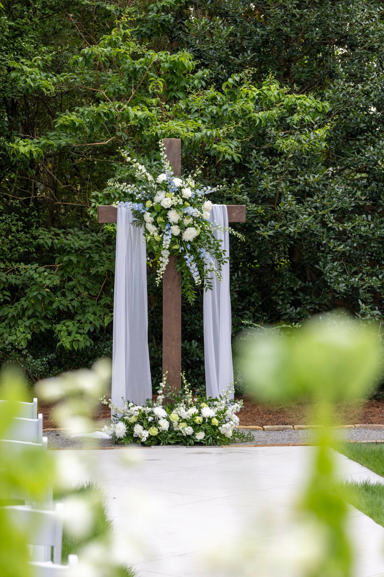 Wooden cross decorated with white flowers and draped fabric, set outdoors among green trees, with a white pathway in front.