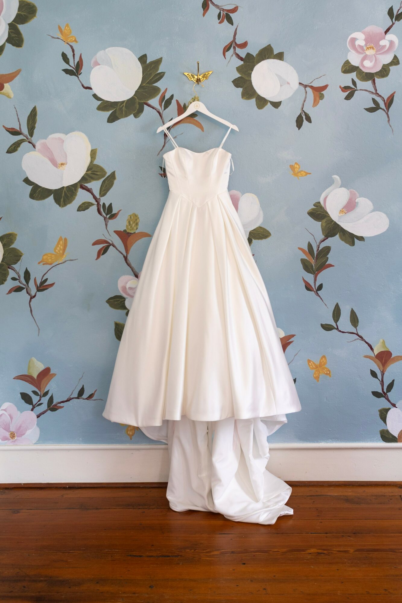White wedding dress hanging on a hanger against a floral wall with swans and flowers, wooden floor below.