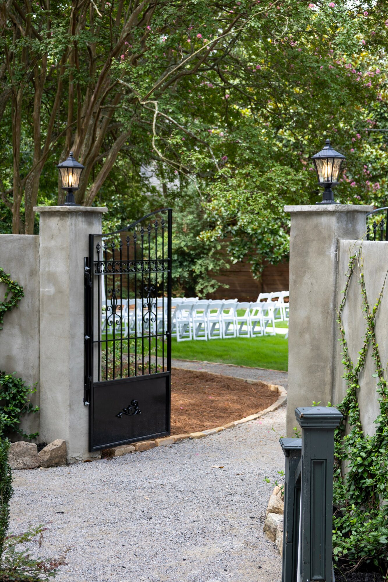 Open black gate with stone pillars and lanterns, leading to a garden with white chairs and green trees.
