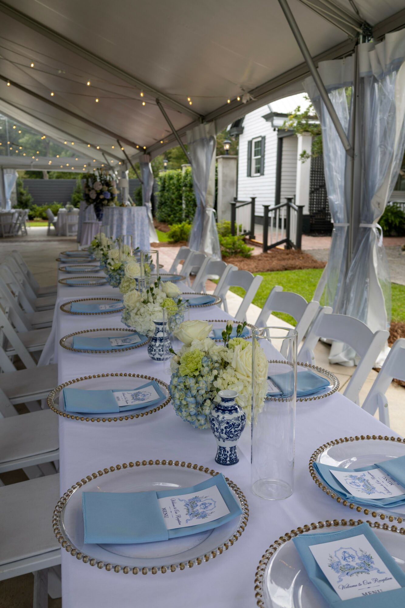 Long outdoor table set with plates, napkins, and floral centerpieces under a tent, with a white building and greenery in background.
