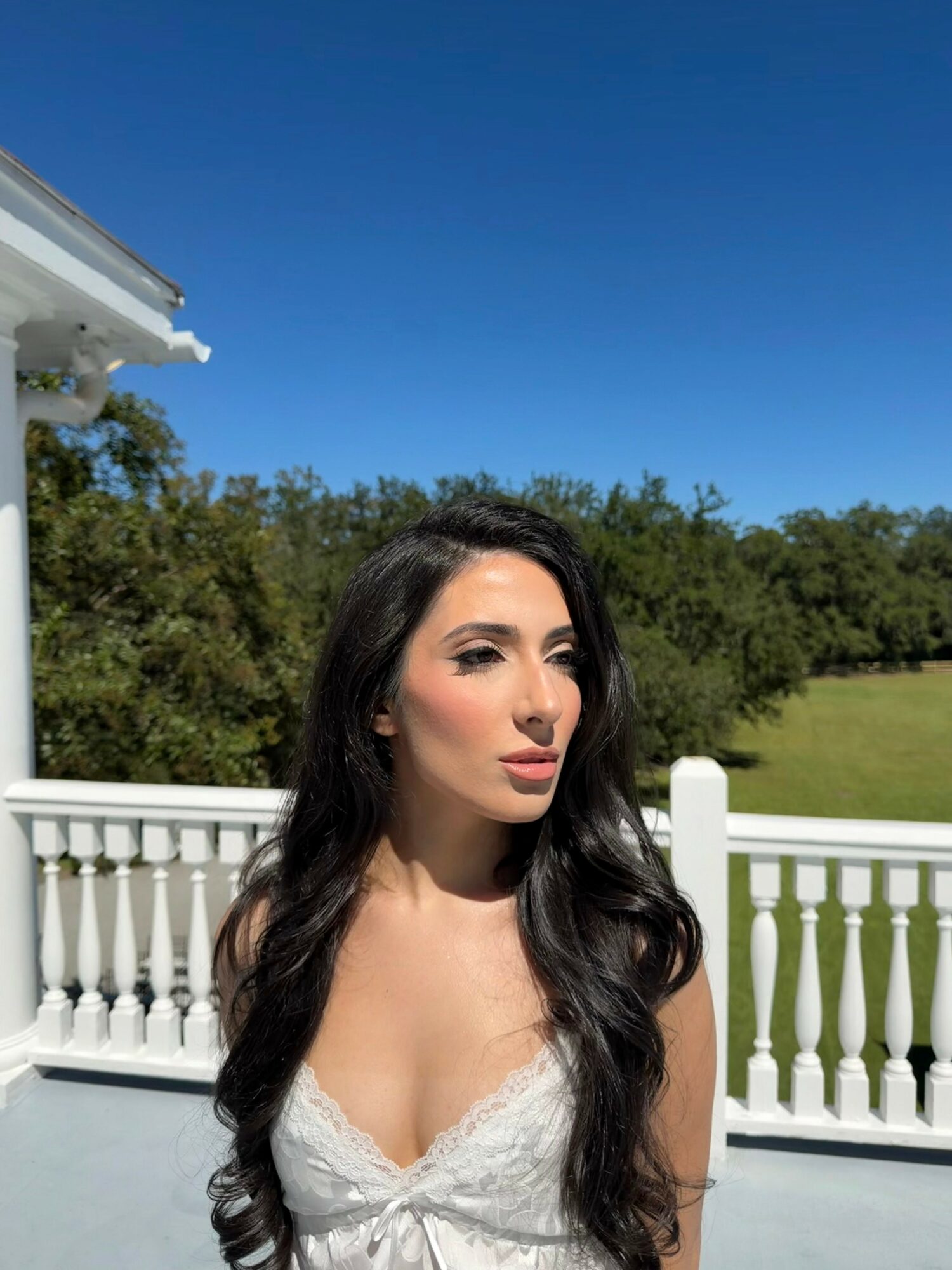 Woman with long dark hair standing outdoors on a balcony with white railing, blue sky, and greenery in background.