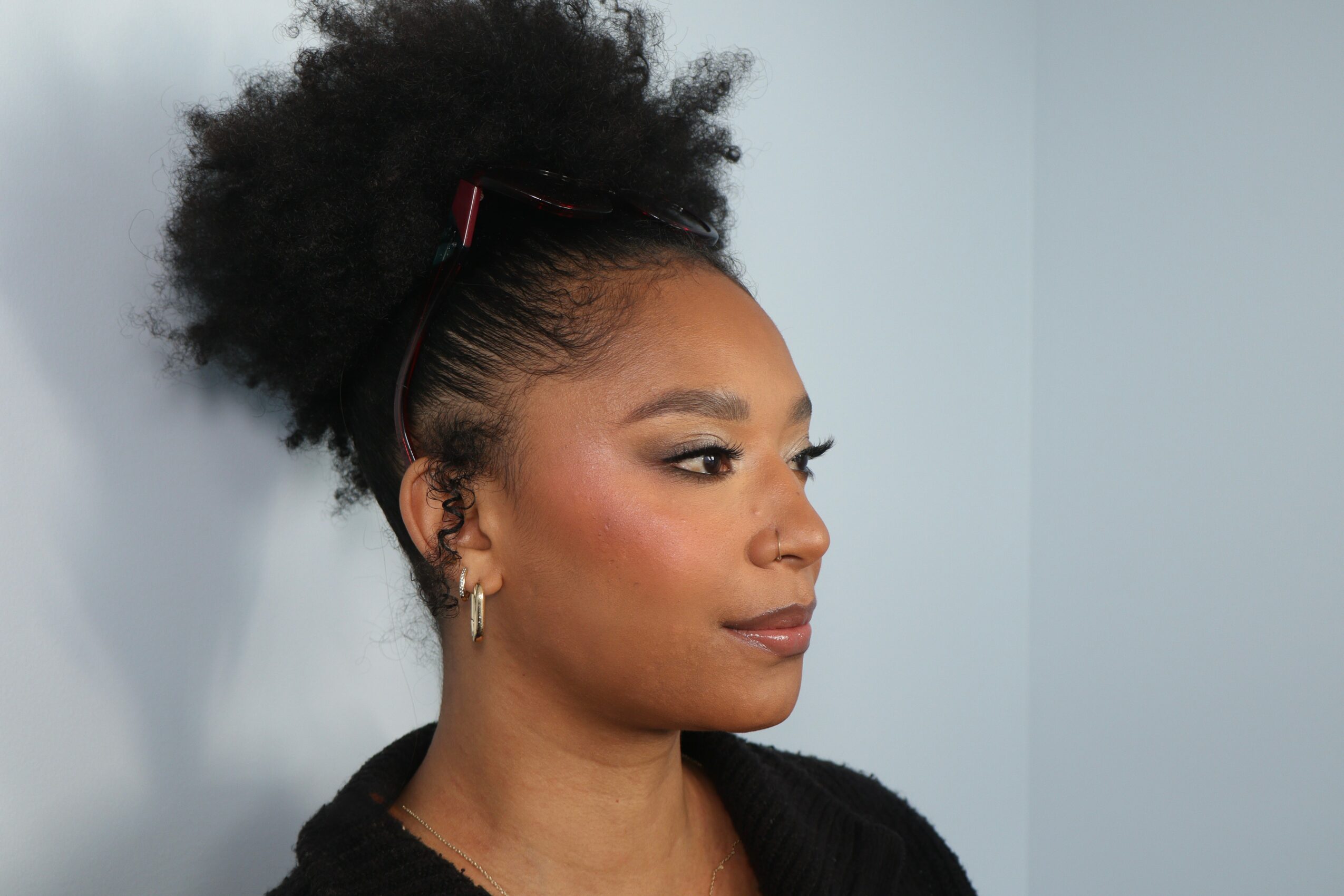 Portrait of a woman with natural hair styled in a puff, wearing earrings and a black top, against a light background.