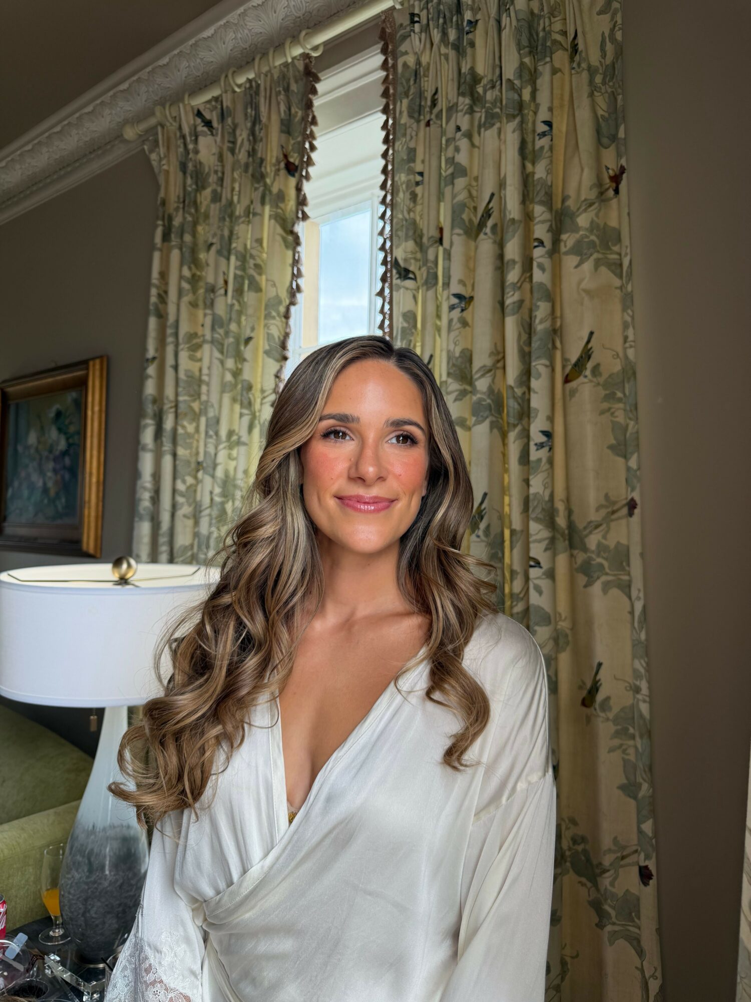 Woman with long wavy hair smiling, wearing a white top, in a room with floral curtains and a painting on the wall.