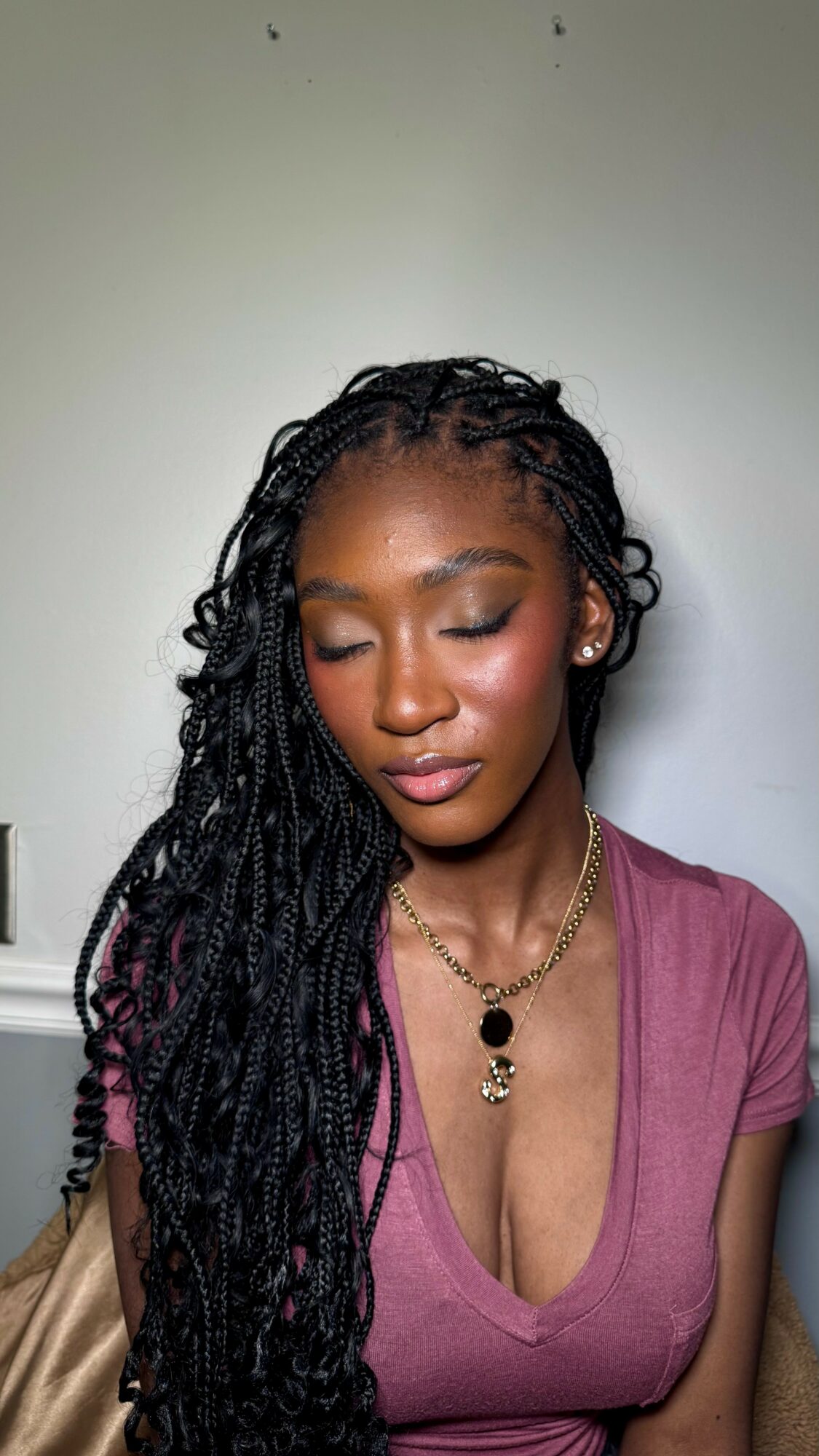 Young woman with long braided hair wearing a pink top and layered necklaces, looking downward.