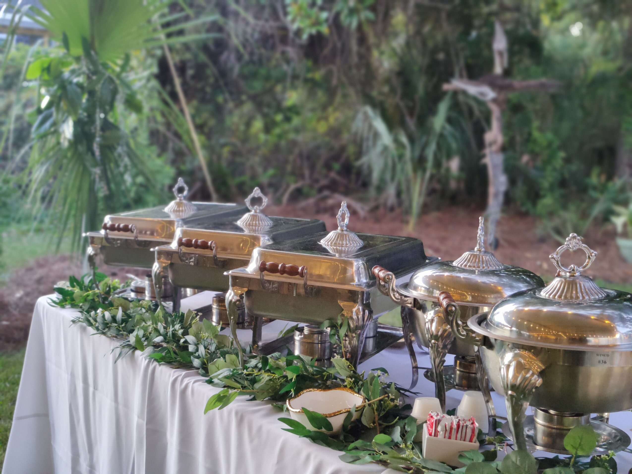 Chafing dishes arranged on a table with greenery and flowers, outdoors with trees and plants in the background.