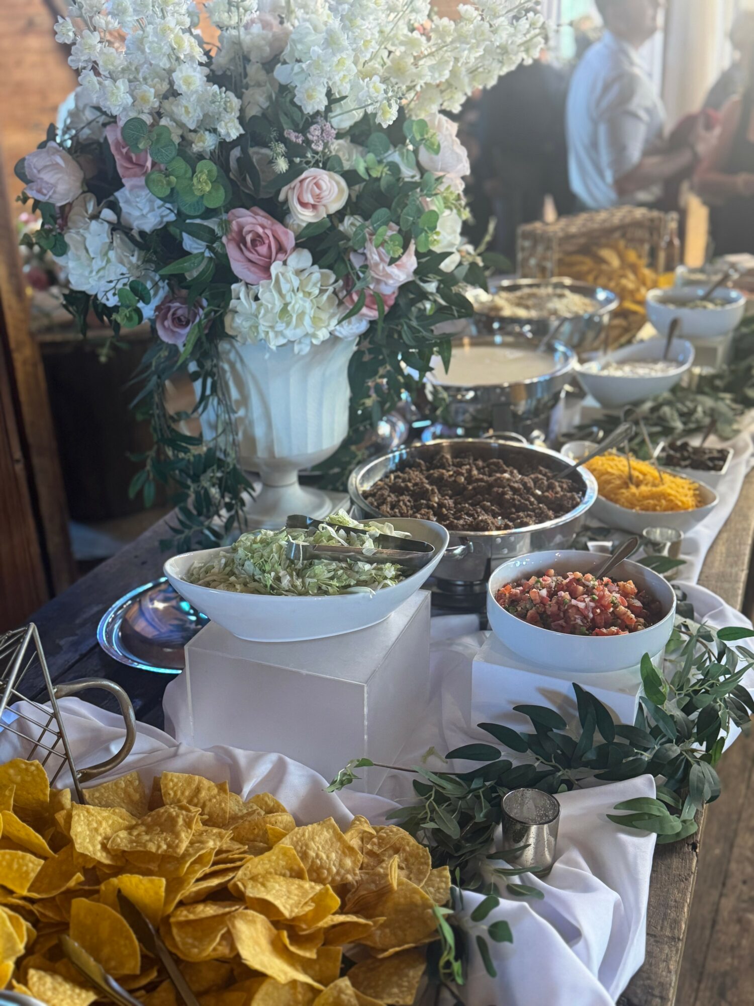 Table with various dishes, a large floral arrangement, and greenery, set for a celebration or event.