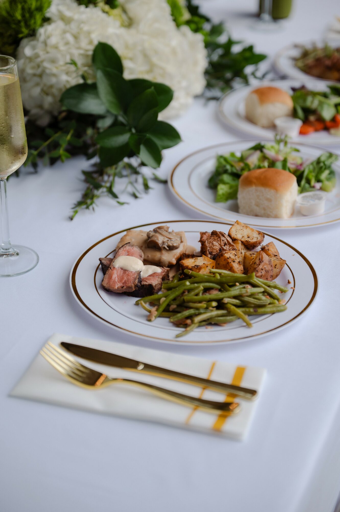 Plate with cooked meat and green beans on a white tablecloth, with a fork and napkin in front, and floral centerpiece in background.