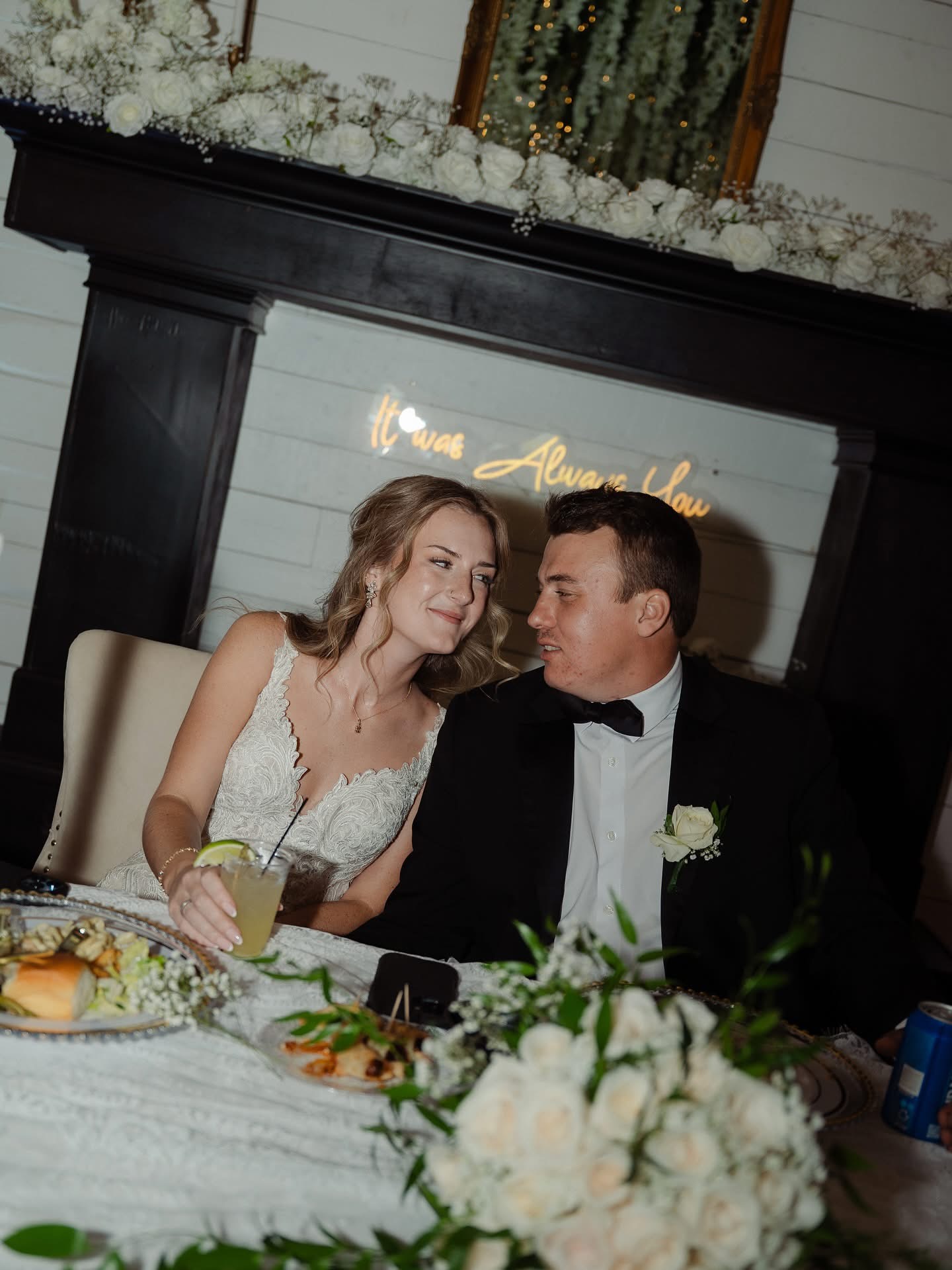 A bride and groom sit close together at a wedding reception, smiling and looking at each other, with a floral arrangement in front.