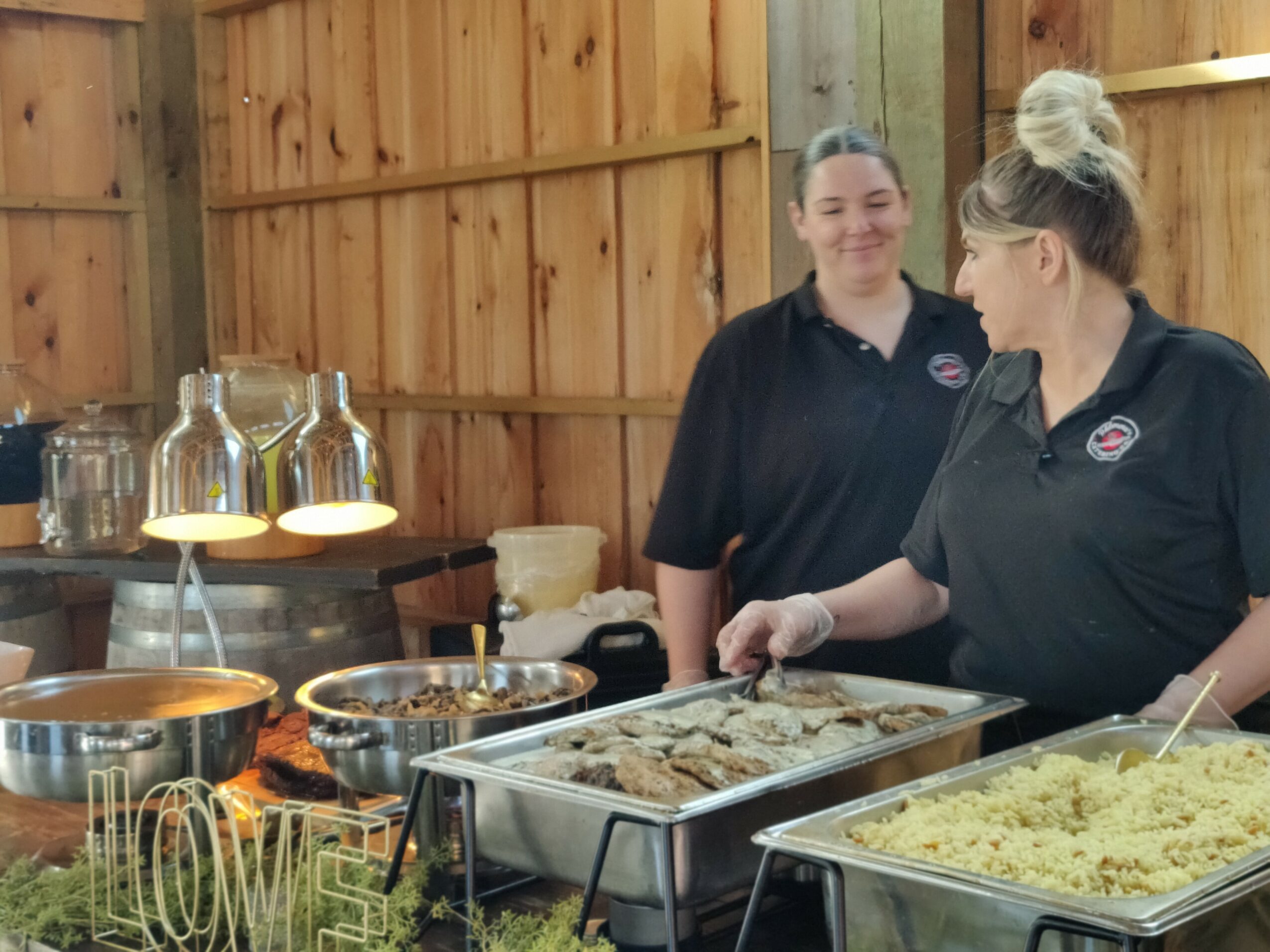 Two women in black shirts preparing food behind a counter with trays of cooked dishes, in a wooden-paneled room.