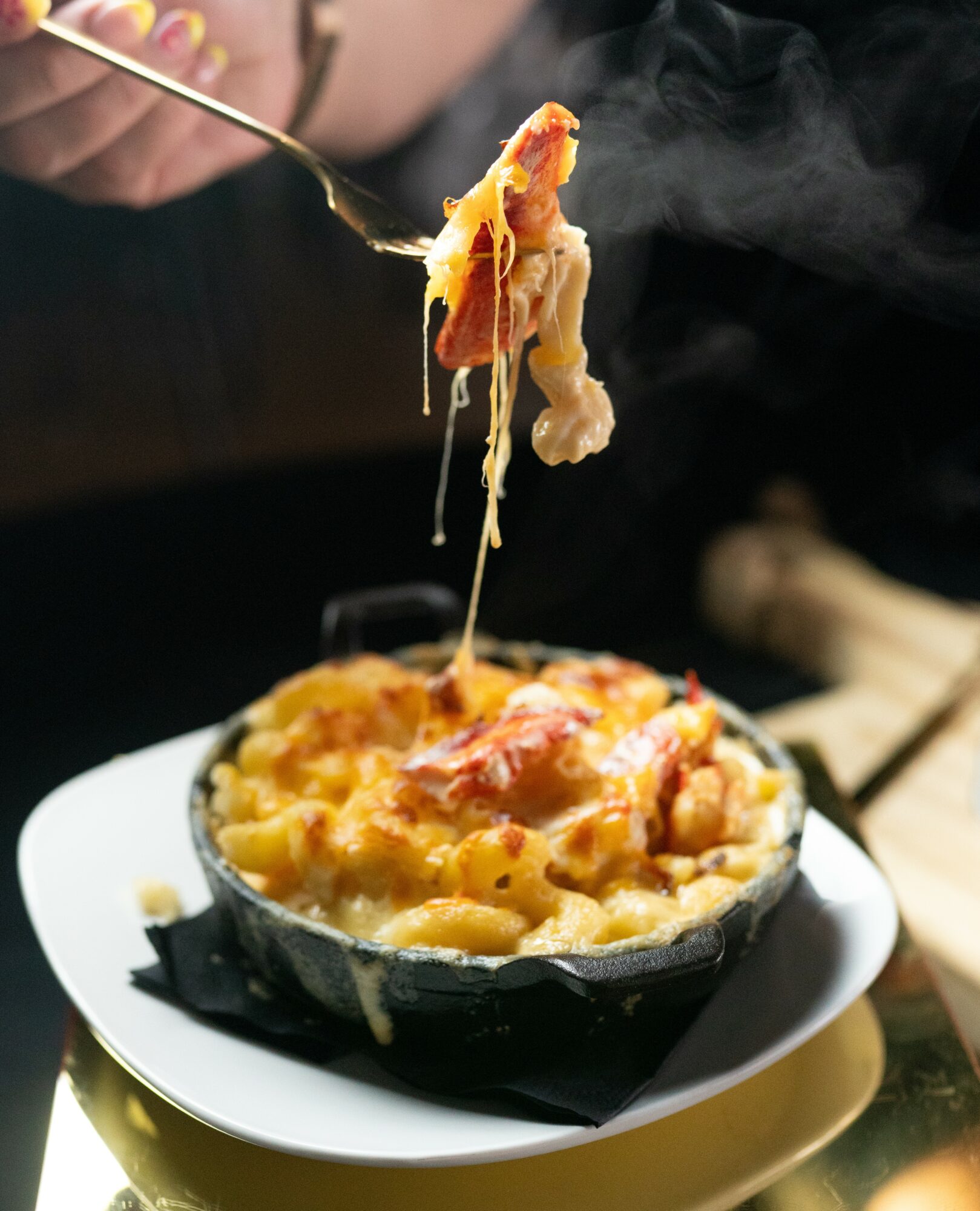 Person lifting a forkful of cheesy baked dish from a black bowl on a white plate, steam rising, with a wooden utensil nearby