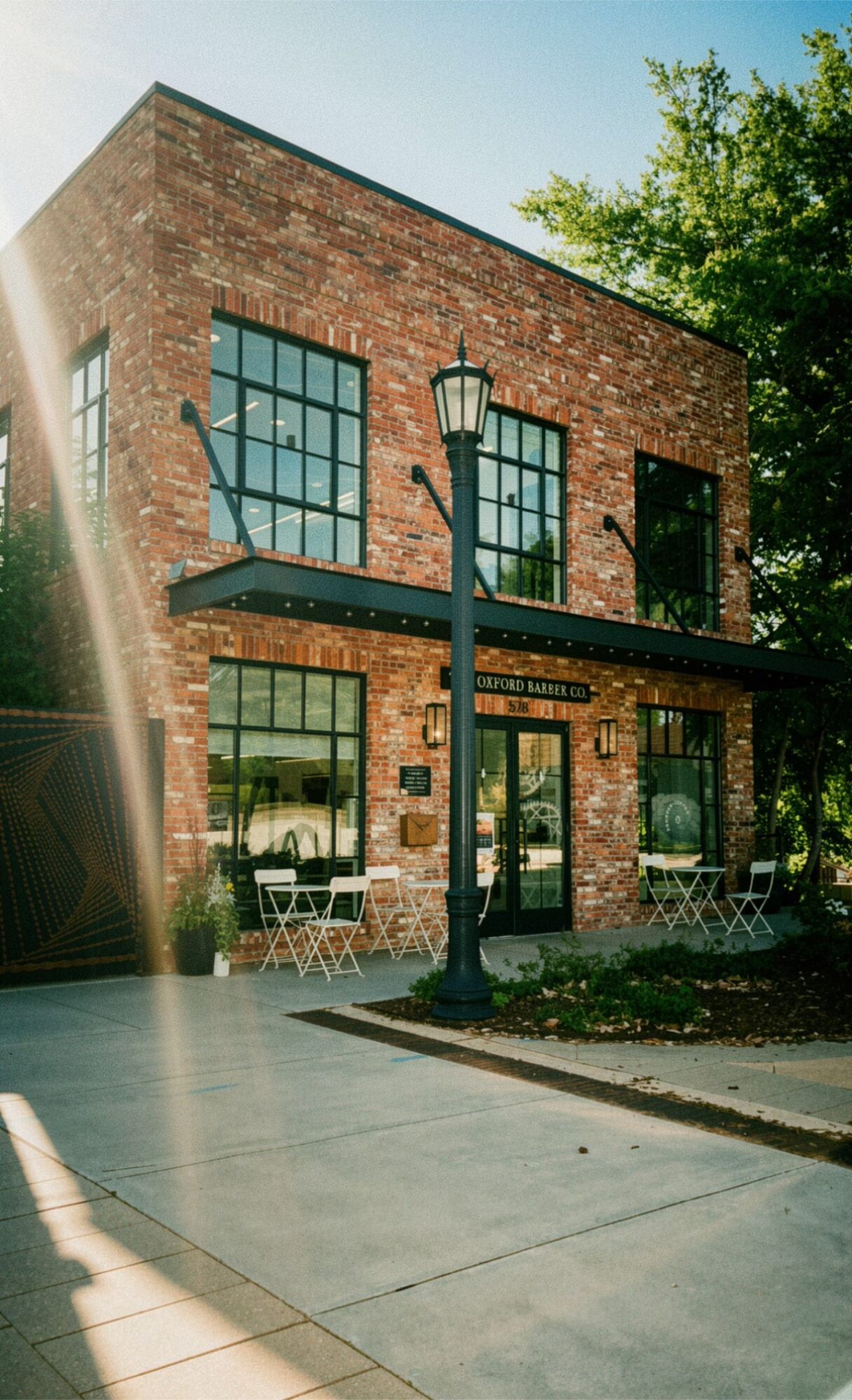 Two-story brick building with large windows, a streetlamp, outdoor café tables, and trees.