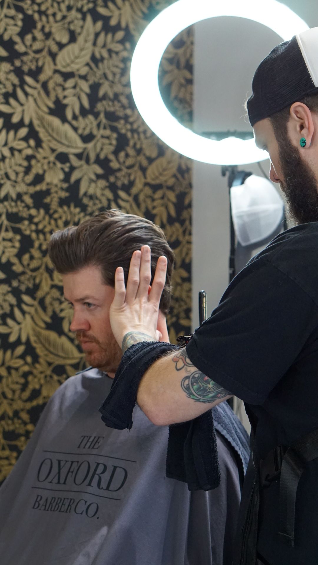 A barber trims a seated person's hair beside a glowing ring light.