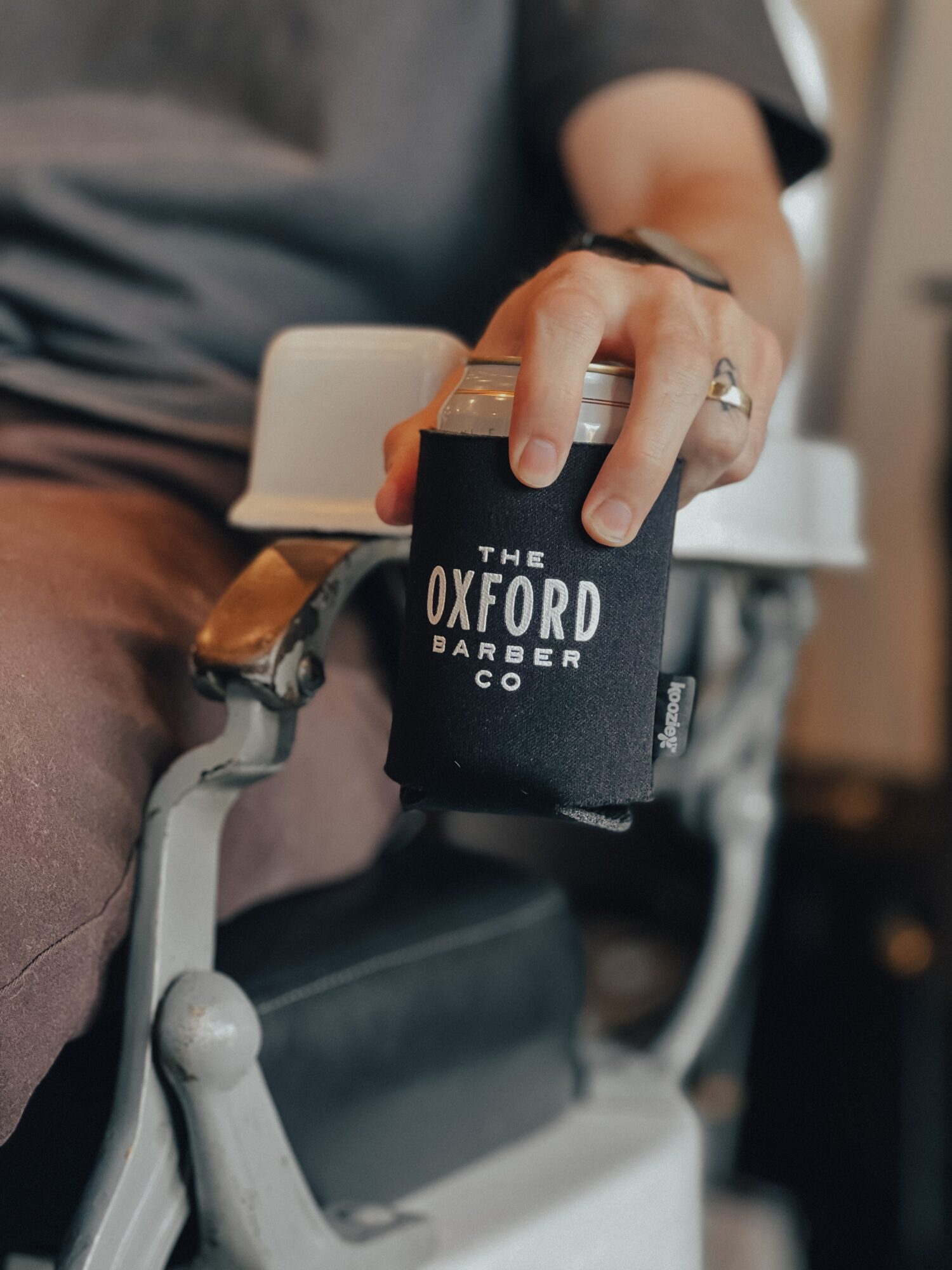 A hand holds a black Oxford Barber Co. mug beside a barber chair.