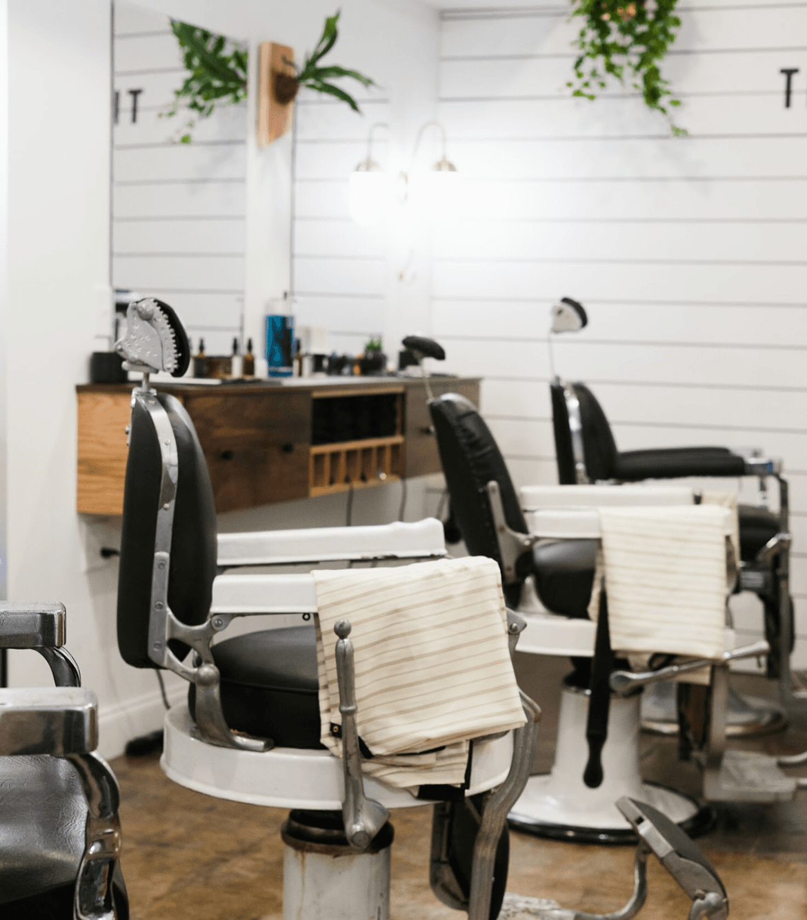 Several black salon chairs with white cushions sit in a bright salon with white paneled walls and a wooden counter.