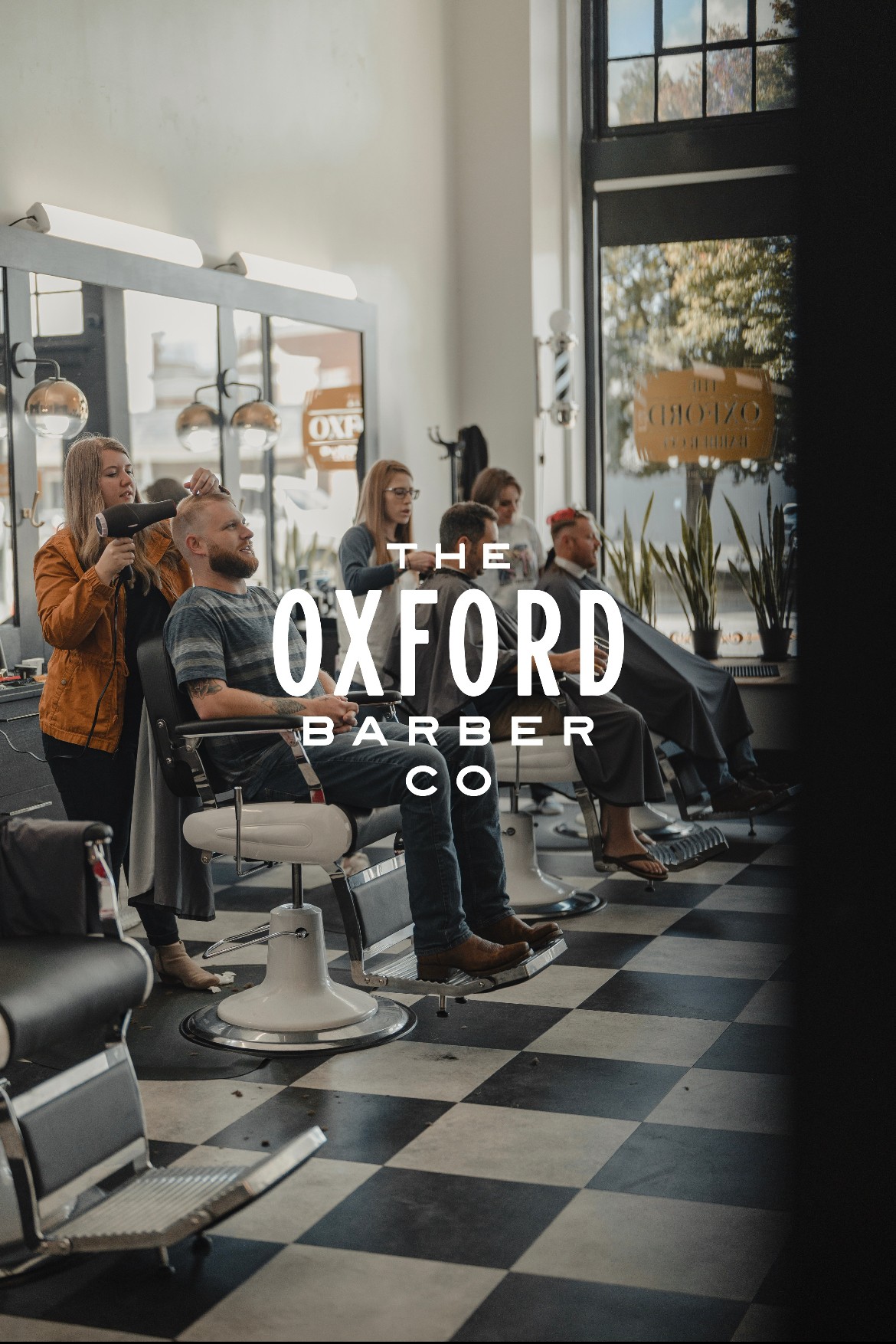 People sit in barber chairs inside a barbershop, with large windows and the words “The Oxford Barber Co.”