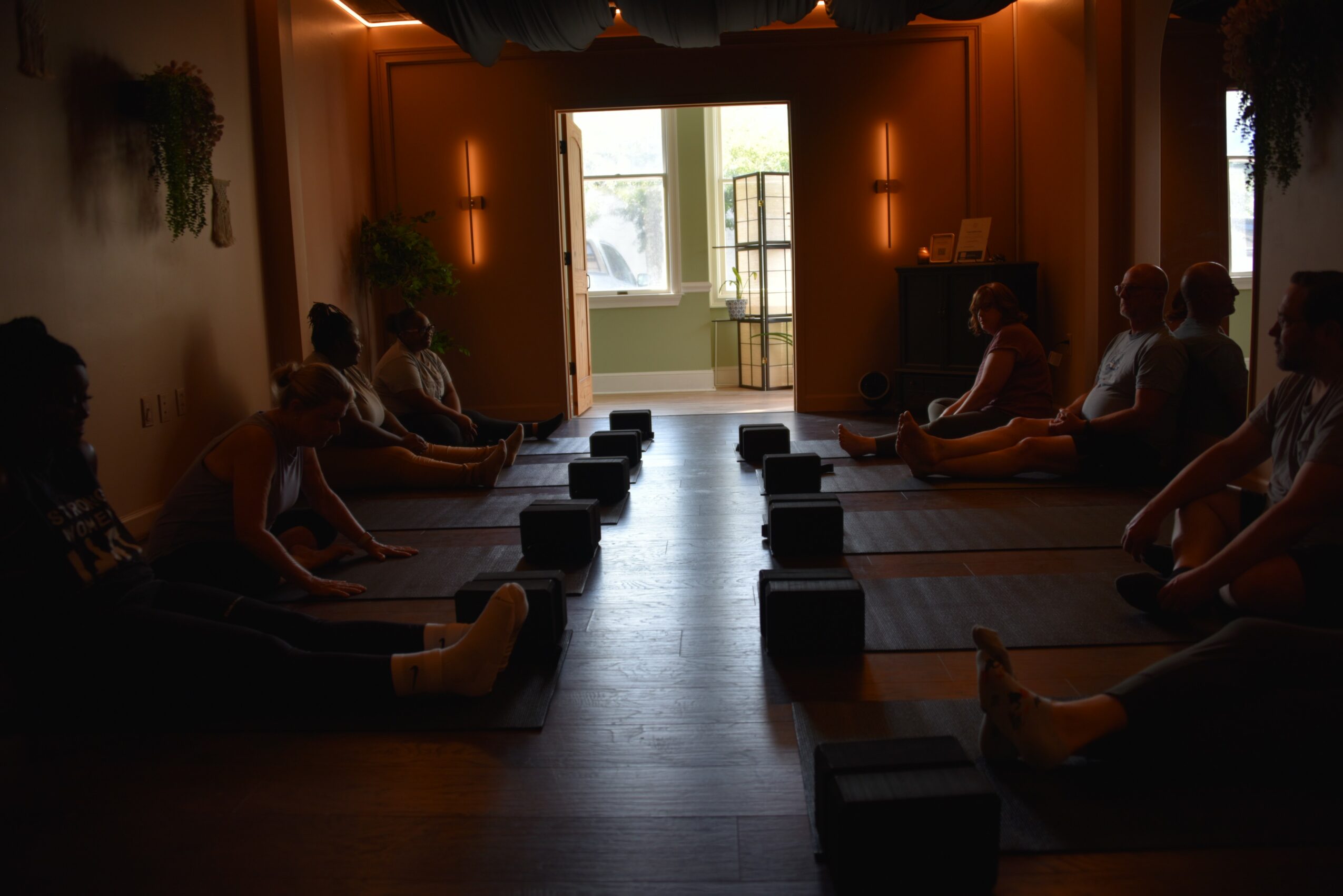 People sitting cross-legged on mats in a dimly lit room, facing a bright window with sunlight streaming in.