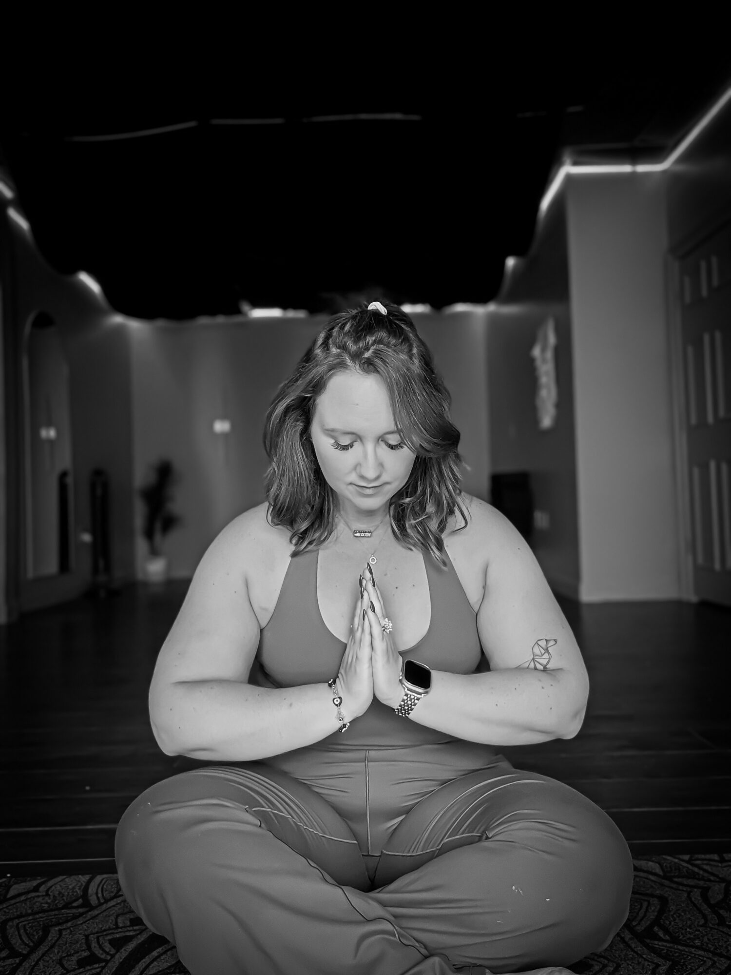 Woman sitting cross-legged with hands in prayer position, eyes closed, in a yoga studio, black and white image.