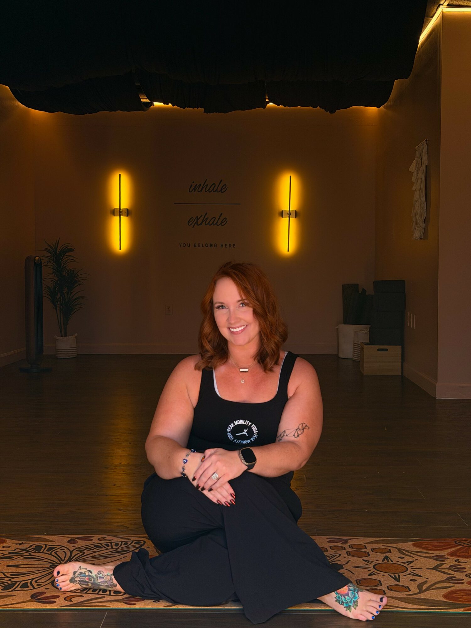 Woman with curly hair sitting cross-legged on a patterned rug, smiling, in a dimly lit room with yellow wall lights.