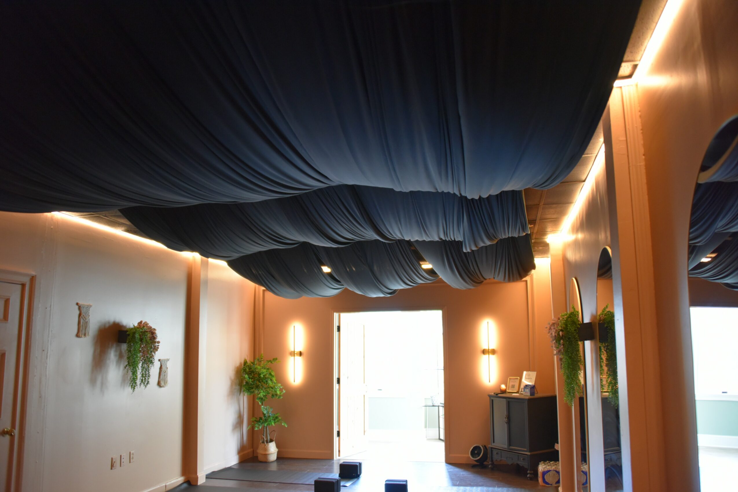 Ceiling with dark blue fabric draped in a circular pattern, illuminated by warm wall lights, with a bright door at the end.