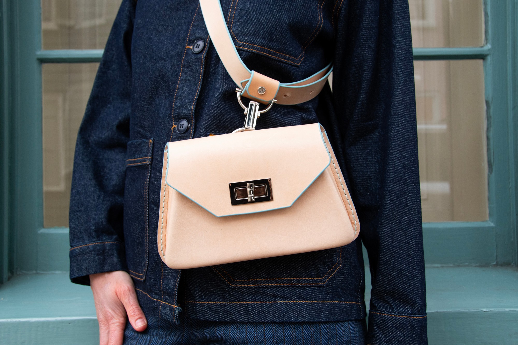 Person wearing a dark denim jacket with a beige handbag hanging from their shoulder, standing in front of a windowed door